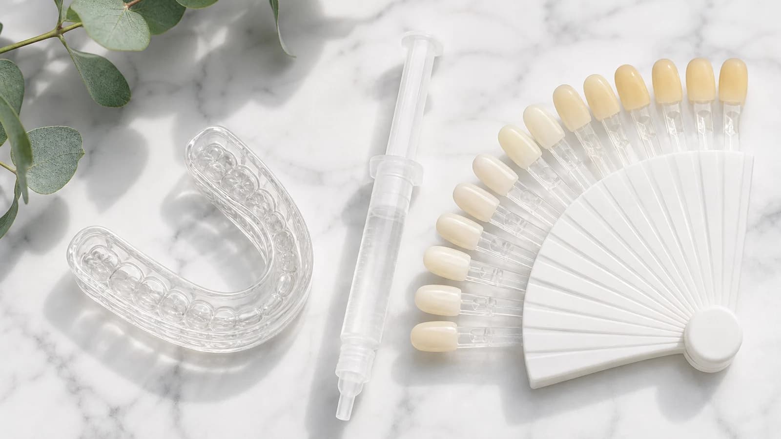A clear at-home whitening tray and gel syringe beside a tooth shade guide on a white marble surface